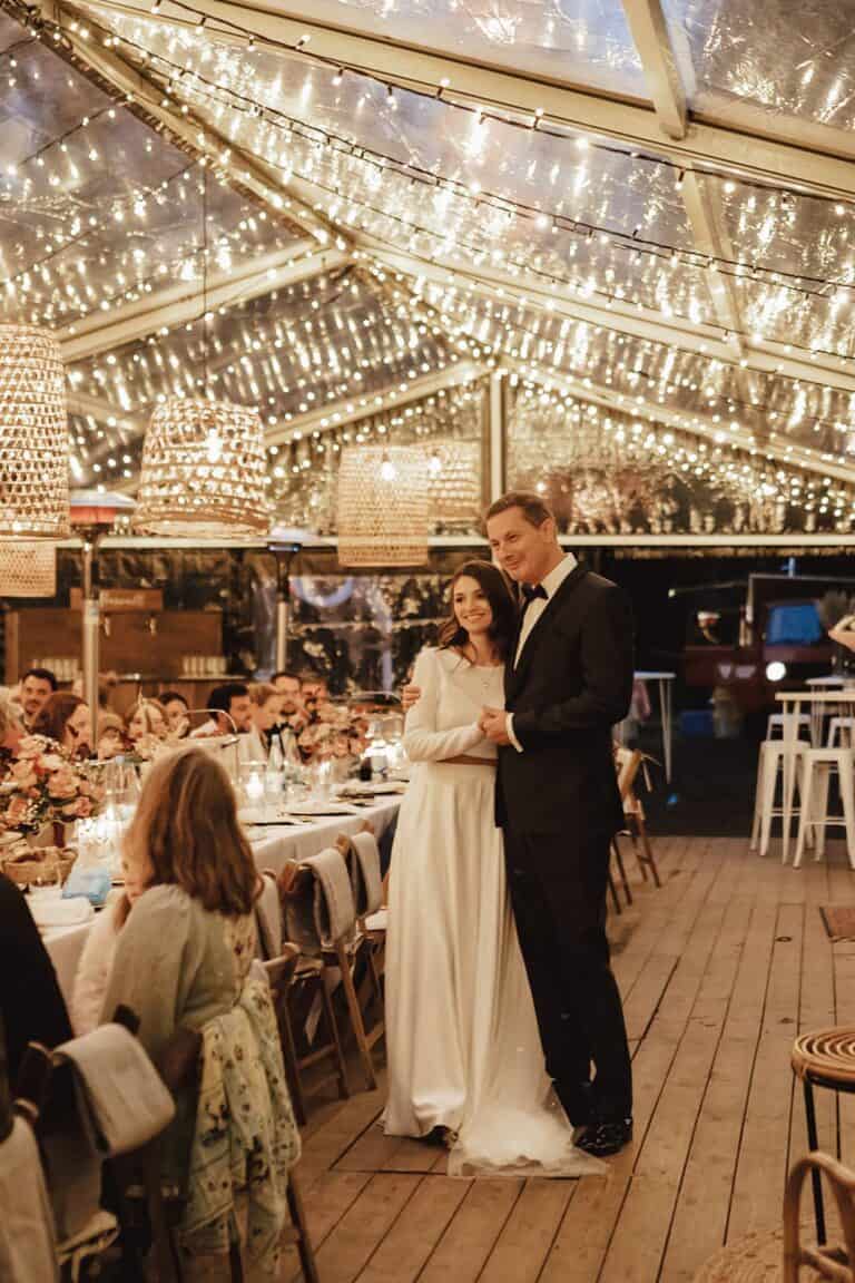 Brautpaar bei einer Outdoor Zelthochzeit geplant vom Hochzeitsplaner in warmer Abendstimmung mit Lichterketten über der langen Tafel
