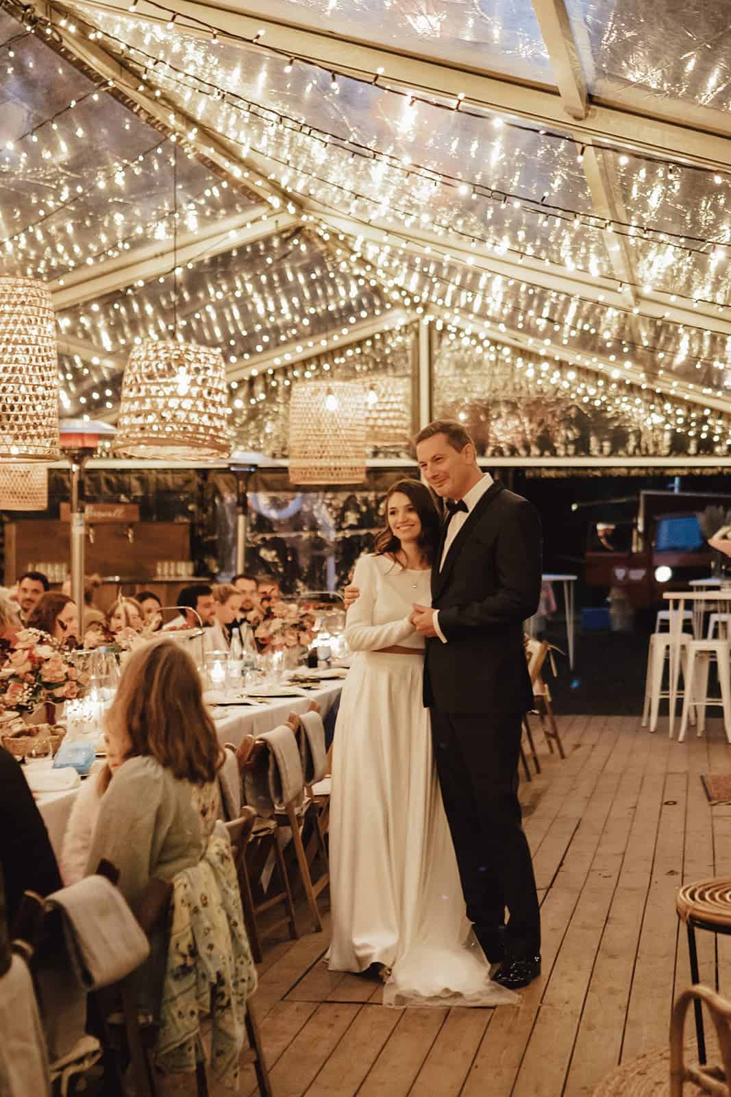 Brautpaar bei einer Outdoor Zelthochzeit geplant vom Hochzeitsplaner in warmer Abendstimmung mit Lichterketten über der langen Tafel