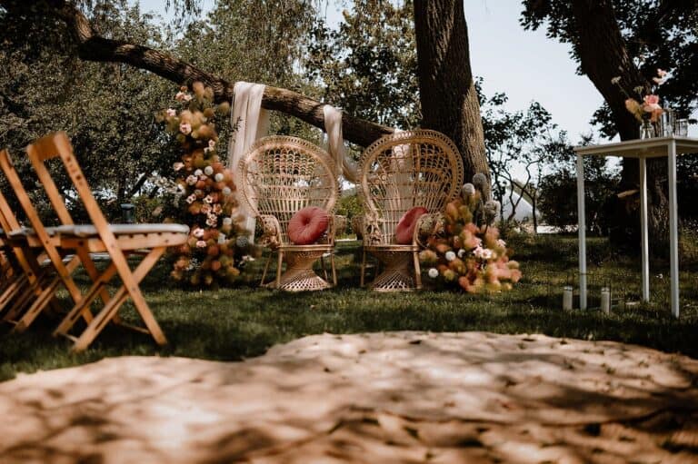 Zeremonie-Setup mit Peacock Chairs, floraler moderner Dekoration und Stoffbahnen für eine exklusive Gartenhochzeit an einem privaten Ferienhaus.