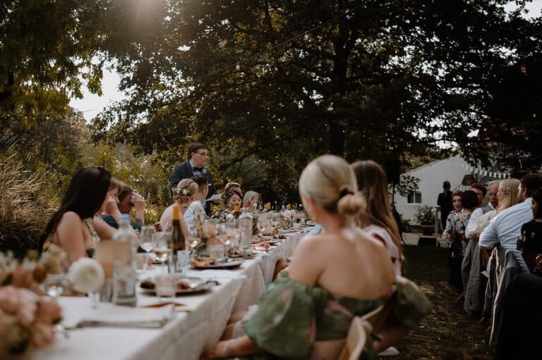 Gemeinsames Hochzeitsdinner mit Reden unter Bäumen und Lichterketten bei einer Outdoor-Hochzeit an einem privaten Ferienhaus.
