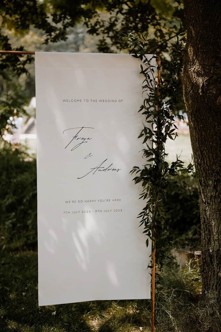 Minimalistisches Welcome-Schild für ein Wedding Weekend an einem Airbnb-Ferienhaus, platziert im Grünen bei einer stilvollen Outdoor-Hochzeit.