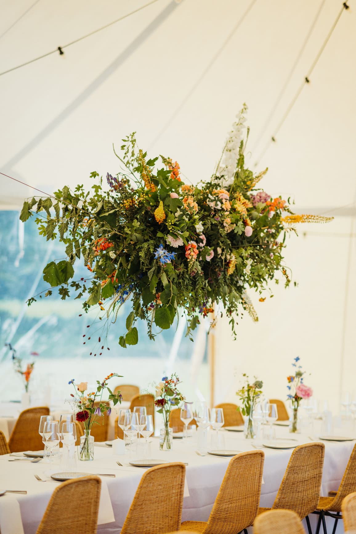 Üppige, schwebende Blumeninstallation über festlich gedeckten Tafeln im Hochzeitszelt – florale Gestaltung einer Gartenhochzeit im eigenen Gartengeplant durch die Hochzeitsplaner von Hey Happy Day.