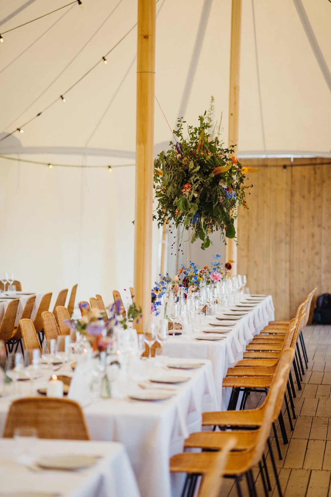Lange, weiß gedeckte Hochzeitstafeln mit Rattanstühlen und farbenfrohen Blumen im Sailcloth-Zelt – individuelle Outdoor-Hochzeit mit Full-Service Hochzeitsplanung.