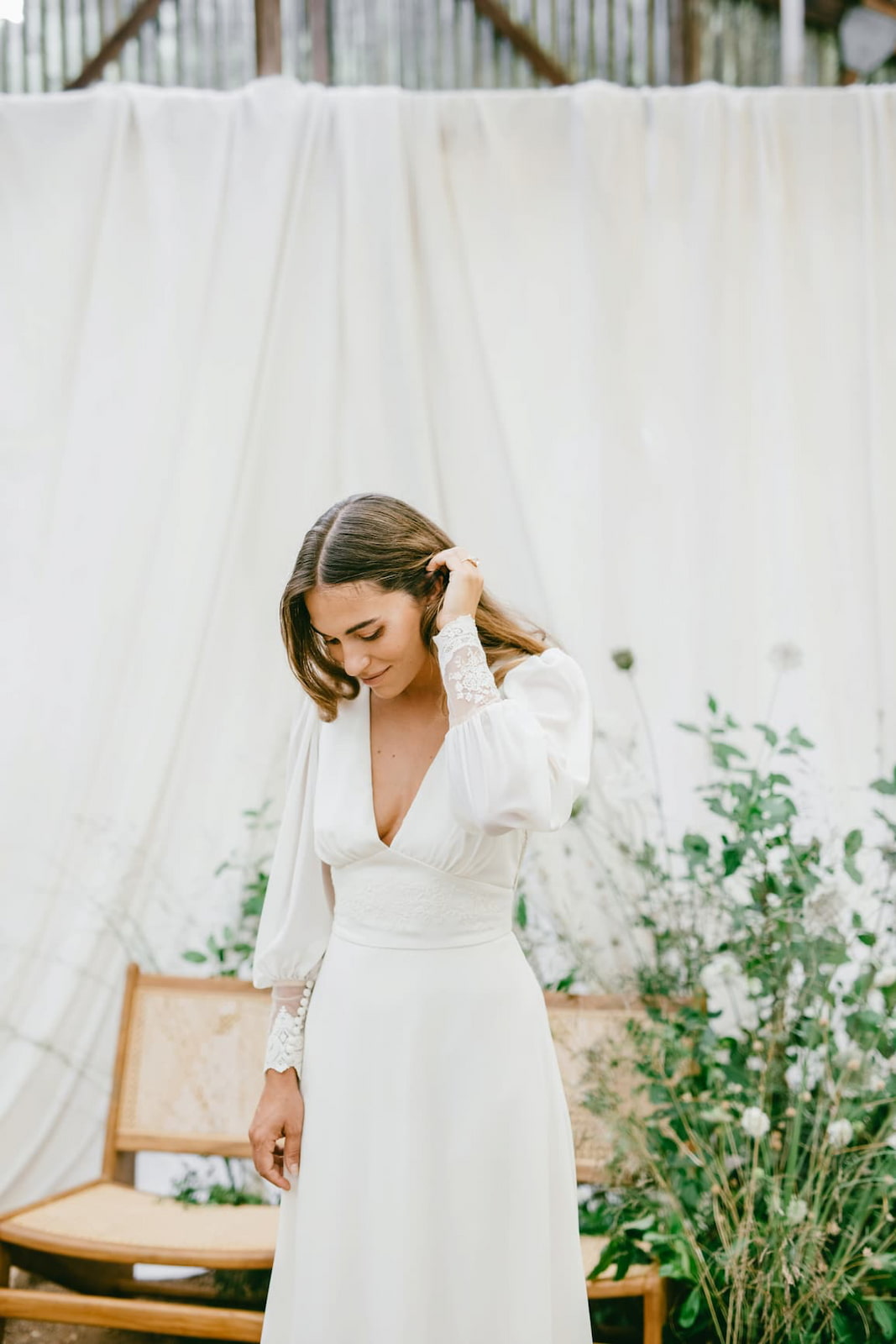 Braut in einem eleganten weißen Hochzeitskleid mit langen Ärmeln vor einem hellen Stoffhintergrund und wilder Floristik – romantische Inspiration für ein Wedding Weekend in Frankreich geplant von den Weddingplannern von Hey Happy Day Hochzeitsplanung und Events.