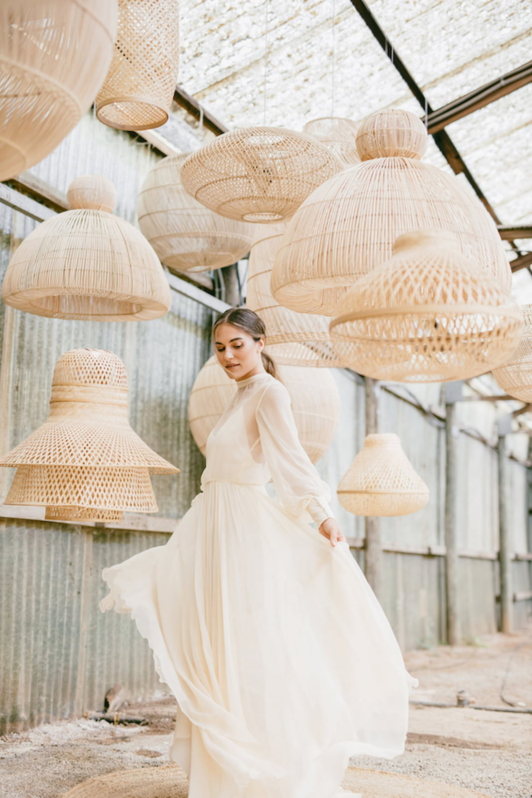 Braut in einem fließenden Hochzeitskleid zwischen großen geflochtenen Rattanleuchten in einer rustikalen Scheune – individuelles Hochzeitsdesign für ein Wedding Weekend mit Full-Service Hochzeitsplanung.