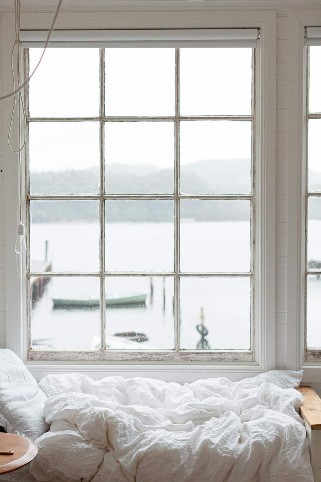 Gemütliches Bett von einem Brautpaar Zimmer in einem Ferienhaus mit weißer Leinenbettwäsche vor einem großen Fenster mit Blick auf einen See – Übernachtung in einer besonderen Unterkunft für ein gemeinsames Hochzeitswochenende.